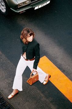 a woman in white pants and black shirt crossing the street with a brown bag on her shoulder