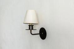 a black wall light with a white shade on it's arm and a lamp attached to the wall