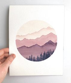 a hand holding up a piece of paper with mountains and trees in the distance on it