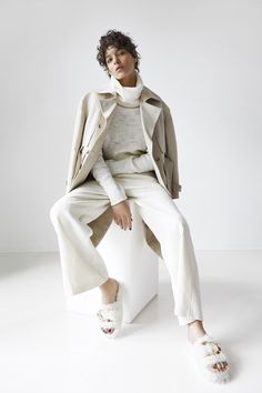 a woman sitting on top of a white block wearing a beige coat and matching pants