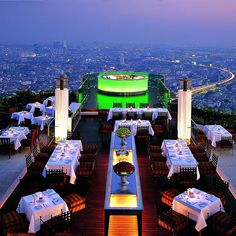 an outdoor dining area with tables and chairs overlooking the city at night, lit up by bright lights