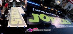 several people are walking around in an indoor area with signs on the floor, and one person is using a skateboard