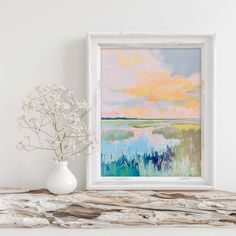 a white vase sitting on top of a wooden table next to a framed art piece