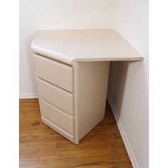 a white desk with three drawers in a corner next to a wall and wooden floor