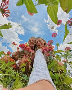 two women are taking a selfie with flowers in the foreground and clouds in the background