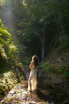 a woman standing in the water next to a waterfall with sunlight streaming through her hair