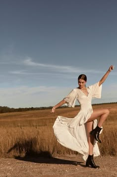 a woman in a white dress and black boots is dancing on a dirt road with tall grass