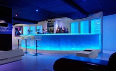 the bar is lit up with blue lights and bottles on the back wall, along with white stools