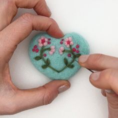 two hands are holding a small blue heart with pink flowers on it and one hand is stitching together