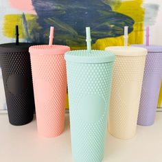 four different colored cups with straws in them sitting on a white table next to a painting