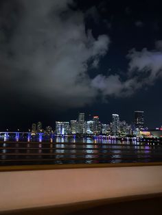 the city skyline is lit up at night as seen from across the water with clouds in the sky