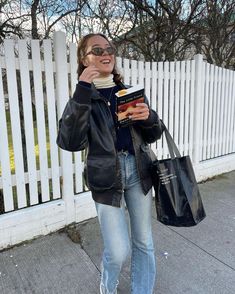 a woman is talking on her cell phone while holding a book and shopping bags in front of a white picket fence