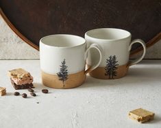 two coffee mugs sitting next to each other on a table with cookies and brownies