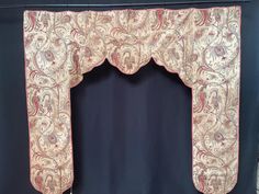 an ornate window curtain with red and white designs on the top, in front of a blue wall