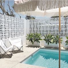 an outdoor pool with lounge chairs, umbrella and trees in the back ground near a white fence