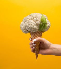 a hand holding an ice cream cone with cauliflower on it, against a yellow background