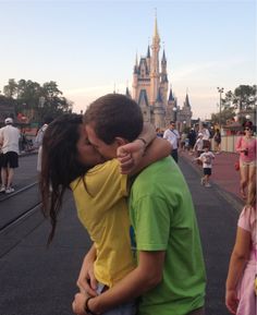 a man and woman kissing in front of a castle with people walking on the sidewalk