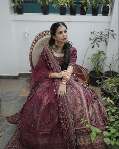 a woman sitting on top of a chair in a purple dress and gold jewelry with potted plants behind her