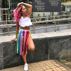 a woman with pink hair wearing a rainbow skirt and white crop top posing for the camera