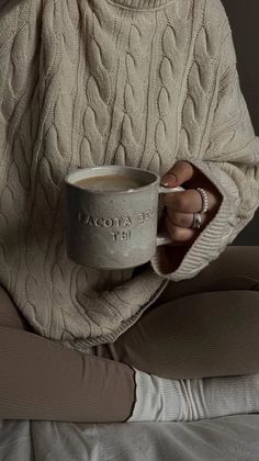 a woman sitting on a bed holding a coffee cup in her hand and wearing a sweater