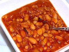 a white square bowl filled with beans and chili sauce on top of a table next to a spoon