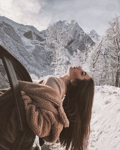 a woman laying on top of a car in the snow next to trees and mountains