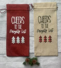 two small bags with words on them sitting next to pine cones and evergreen needles, one is red and the other has white writing that says cheers to the