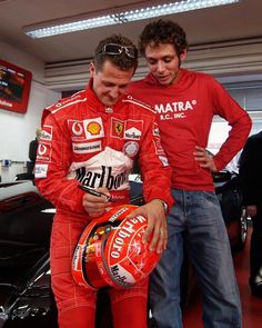 two men standing next to each other in front of a car with a helmet on