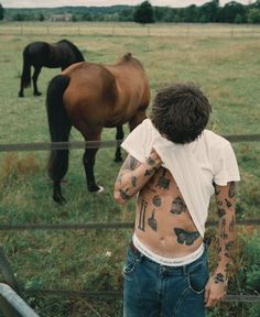 a man with tattoos on his back standing in front of a fence next to two horses