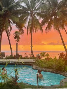 two people are swimming in an outdoor pool with palm trees and the sun setting behind them