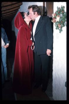 a man and woman dressed in red kissing each other while standing next to a christmas wreath