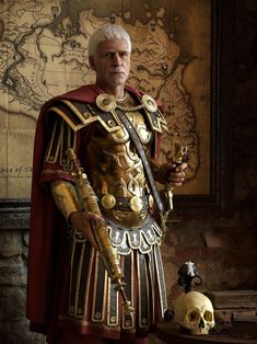 a man dressed in armor standing next to a table with a skull on it and a map behind him