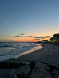 the sun is setting at the beach with rocks in front of it and buildings on the other side