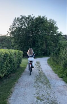 a woman riding a bike down a dirt road next to a lush green field and trees