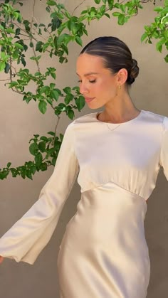 a woman in a white dress standing next to a tree with green leaves on it