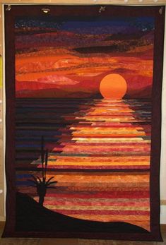 a quilted wall hanging with the sun setting over water and cactus in foreground