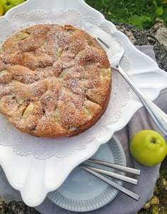 an apple pie is on a white platter with silverware and green apples in the background