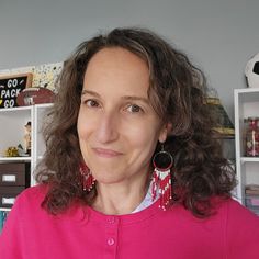 a woman with curly hair wearing a pink shirt in front of bookshelves and shelves