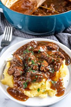 a white plate topped with mashed potatoes covered in beef and gravy next to a pot of stew