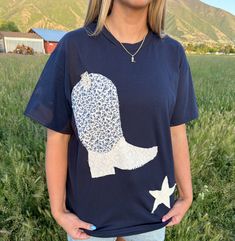 a woman standing in a field wearing a t - shirt with a cowboy boot on it