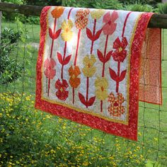 a quilt hanging on a wire fence in a field with flowers and grass behind it