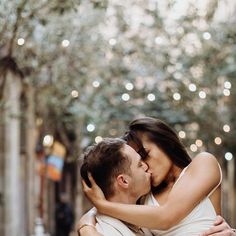 a man and woman kissing in an alleyway with lights on the trees behind them