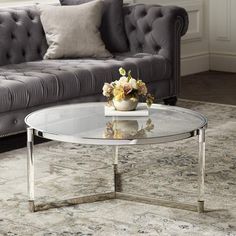 a glass coffee table with flowers on it in front of a gray couch and rug