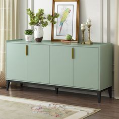 a green cabinet with two plants on top and a framed picture above it in front of a window