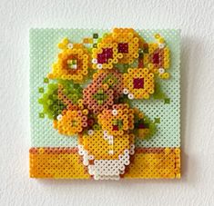 a cross - stitch flower arrangement on a white wall hanging from a peg board in the shape of a vase with yellow flowers