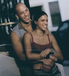 a man and woman sitting next to each other in front of a gate smiling at the camera