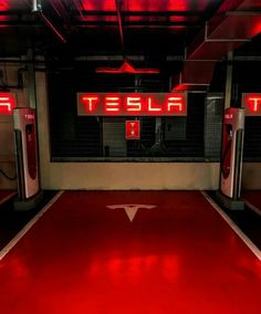 a red carpeted parking garage with two tesla charging stations in the center and neon signs above them