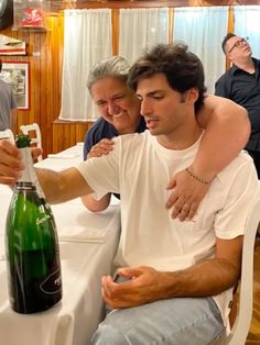 two men sitting at a table with a bottle of wine in front of them and people standing around