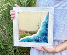 a woman holding up a framed painting with an ocean wave in it's reflection