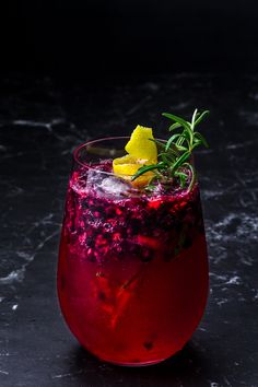 a red drink with lemons and rosemary garnish in a glass on a black surface